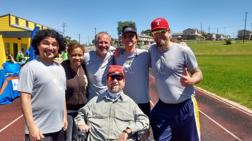 On an outdoor track, a group of smiling people of various ages stand shoulder to shoulder; a man in a power chair is in front.
