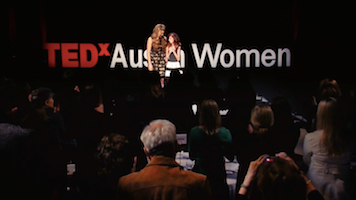 From the behind a full audience, two women stand on a stage with a TEDx Austin Women backdrop. As they look at each other, one has her arm around the other's shoulders. The other crosses her hands over her heart.