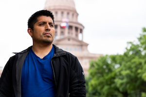 A man looks somberly past the camera, with the Texas State Capitol out of focus in the background