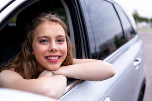 A young woman leans on her folded arms out of a car window and smiles at the camera.