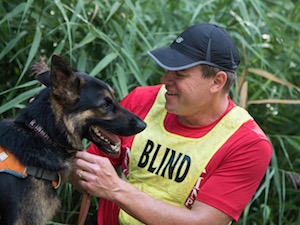 Amid tall blades of grass, a smiling man with a cap and yellow vest that reads Blind adjusts the collar of a panting German Shepherd.