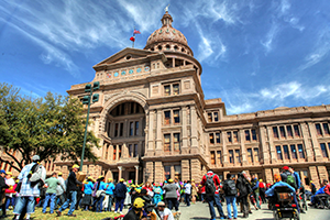 A crowd of people, many wearing yellow caps, stand or sit in front of the Texas Capitol, which stretches in a bright blue sky.