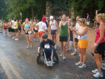 A man using a racing wheelchair passes a line of race event spectators. A woman walks along side him.