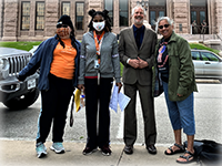 Four people in jackets pose in a line on a sidewalk outside the Texas Capitol.