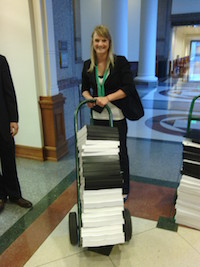 A young woman in business dress holds in both hands the handle of a cart full of bound volumes and smiles directly at the camera.