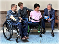 On a wooden bench, a man in a blue suit flips through a booklet. Next to him, with thier attention on the booklet are a smiling young man with no arms or legs in a wheelchair, a pensive young man sitting on the other side of the bench, and a man in a wheelchair with glasses.