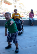 A little boy laughs and reaches his hand toward the camera while a dancer sitting in a wheelchair and spectators sitting farther back look on and smile.