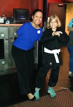 A woman with no arms and girl in a black karate uniform stand side by side, grinning and touching the toes of their right feet together.