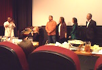 Shot from over the tops of theater seats, four people stand in front of a blank movie screen, all looking at another person at the side of the audience who is gesturing with both hands.