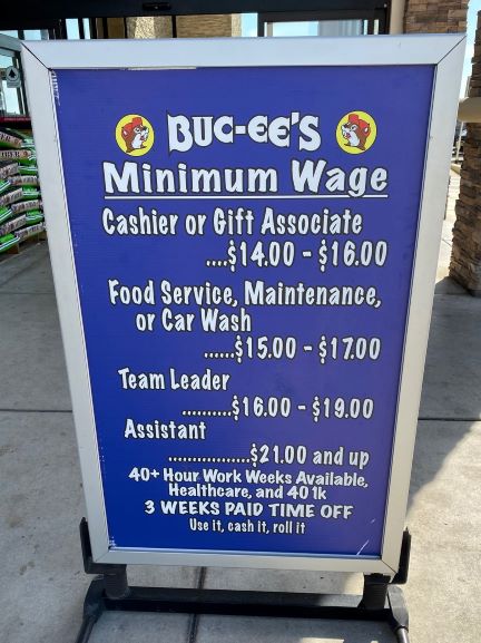 Blue sign advertising Buc-ee's Minimum Wages: Cashier or Gift Associate $14.00-$16.00. Food Service, Maintenance or Car Wash $15.00-$17.00. Team Leader $16.00-$19.00. Assistant $21.00 and up. 40+ Hour work weeks available, healthcare, and 401k. 3 weeks paid time off. Use it, cash, roll it.