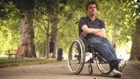Under the shade of trees, a man sits back in his wheelchair, crossing his ankles and arms, with an unamused expression on his face.