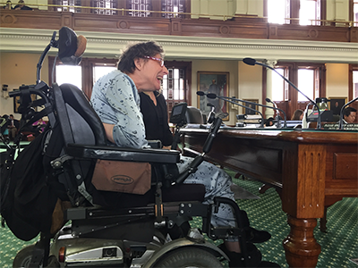 In profile, Susie leans forward in her power chair to speak into a microphone in a large, stately room.