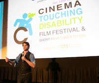 RJ stands in front of a theater screen with the Film Fest logo projected onto it. He holds a mic in one hand and gestures with the other, as if he's making a point.