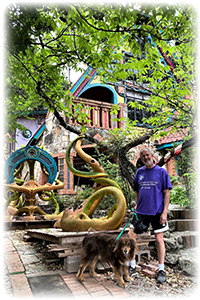 Dennis and his Australian shepherd Kip stand outside a large orange and turquoise house and a set of round concrete and mosaic steps. Trees and swirling gold sculptures share the foreground.