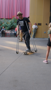 A man in a black t-shirt and fedora glides around a paved area on a skateboard, with a crutch under each arms