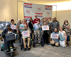 A group of about 20 people with various disabilities stand, sit, and kneel in front of a REV UP banner, some holding signs that read Vote!