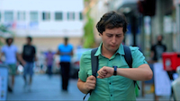 A young man with a worried expression checks his watch. The camera is focused on him while other people and buildings are out of focus in the background.
