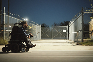 At dusk, a man in a powerchair speeds past a chainlink fence.