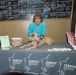 On a display table with brochures and a basket, a woman smiles broadly and leans over a sandy colored dog, who is looking to the right.