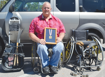 A man sitting in a push wheelchair holds a framed medal in his lap. He sits in front of a grey van and is surrounded by adaptive spots equipment, including sled hockey gear and a racing wheelchair.