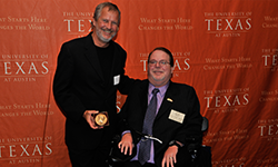 Two men, one standing, one seated in the power chair, show off a golden award.