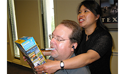 A woman holds a Cheerios box up to the face of a man, who holds a pen in his mouth to sign it.