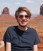 A man in sunglasses smiles at the camera with a sweeping desert landscape in the background.
