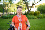 On a green lawn with blurred trees and a house in the background, a woman in a bright orange shirt poses in her power chair and smiles at the camera.