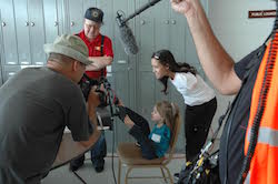 A film crew surrounds a young girl in profile with no arms, sitting back in a chair, feet to an airplane steering device. Jessica Cox, also without arms, standing, bends down behind her and smiles.
