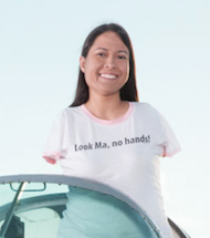 Jessica Cox, a young woman with no arms, stands up behind a windshield. She wears a shirt that reads, Look Ma, no hands!