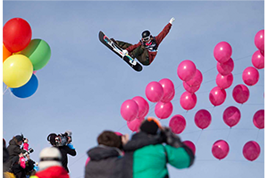A snowboarder soars over a crowd of photographers and colorful balloons.