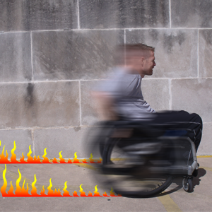 Viewed in profile, a man races by in a wheelchair, blurred and leaving a trail of flames.
