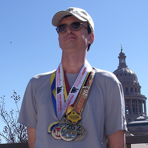 A man in a baseball cap and sunglasses gazes past the camera. He wears about 15 racing medals around his neck.
