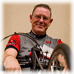 A man with glasses and an athletic t-shirt smiles down at the camera, with his hand-cycle wheel out of focus at the bottom of the frame.