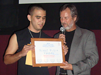 An older man speaking into a microphone hands a certificate to a younger man whose eyes are closed.