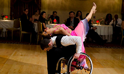 A man in a tuxedo bends forward in his wheelchair while a woman in a formal gown lays on his back with a leg in the air.