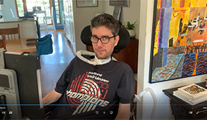 Screenshot of Ady sitting in his power chair, looking meaningfully out at the camera. He has a ventilator tube at his throat and his speaking device is visible in the foreground.
