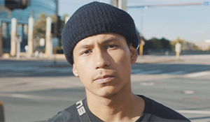 Close up of Brian, in a black beanie and black shirt, looking into the camera with a slightly furrowed brow. A blurred street corner is in the background.