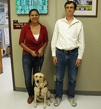 A young woman with a blond guide dog and a tall man smile at the camera