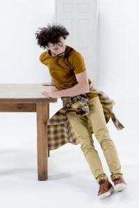 In a stark white room, a man leans all of his weight on his hands on a brown table. His face is calm, but his hair and a shirt tied around his waist are in the air, as though he's moving quickly.