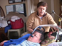 A woman in a bed looks at a breathing tube on her face, while another woman behind the bed adjusts it.